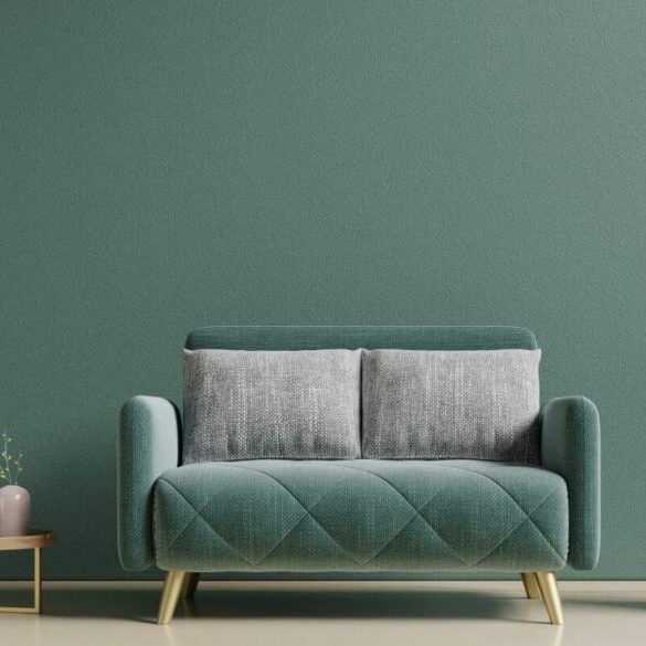 A green sofa with two gray cushions is set against a green wall. On either side are round tables, one with plants and books, the other with decorative vases. Three gold pendant lights hang above.